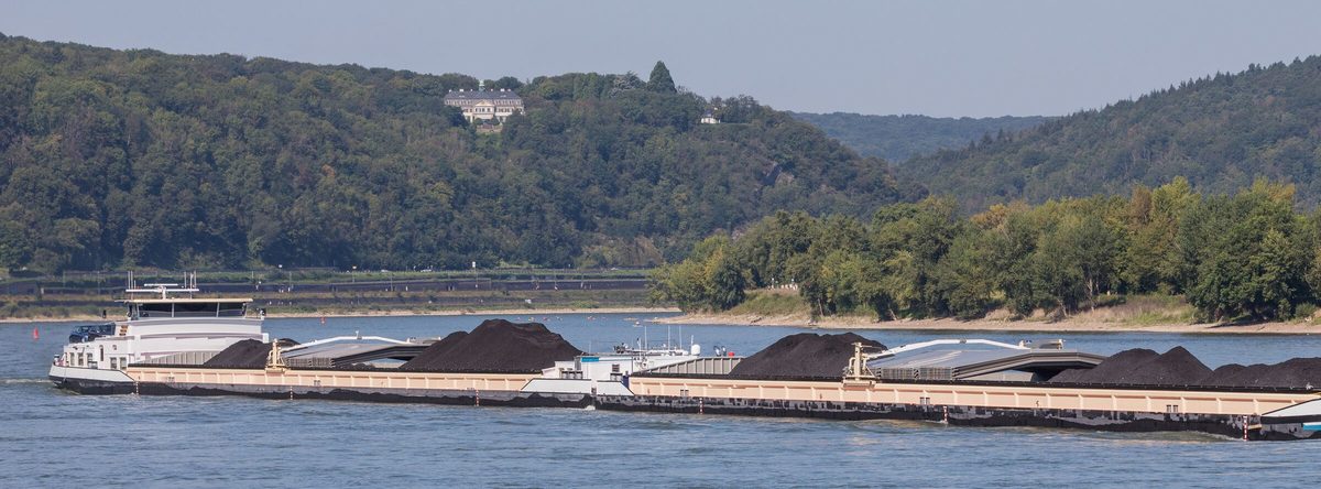 Een lang koppelverband vol steenkool vaart op een brede rivier, met beboste heuvels op de achtergrond